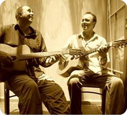 A sepia-toned or filtered photo of two men seated and smiling while playing acoustic guitars.